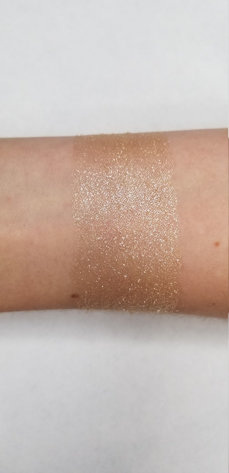May include: A swatch of shimmering, golden-bronze eyeshadow applied to the inner forearm. The product has a high shimmer finish, reflecting light. The skin tone is fair, providing a contrast to the product.
