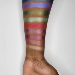 Puede incluir: Un primer plano de un brazo con muestras de sombra de ojos en varios colores, incluyendo azul, verde, rojo, morado y dorado.