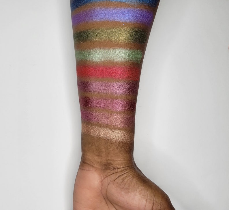 Puede incluir: Un primer plano de un brazo con muestras de sombra de ojos en varios colores, incluyendo azul, verde, rojo, morado y dorado.
