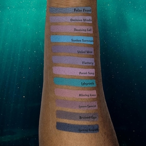 May include: A series of eyeshadow swatches in various shades of blue, purple, and pink are displayed on an arm. Each swatch has a name printed above it, including "False Front," "Ominous Winds," and "Sunken Sorrows." The background is a teal gradient.