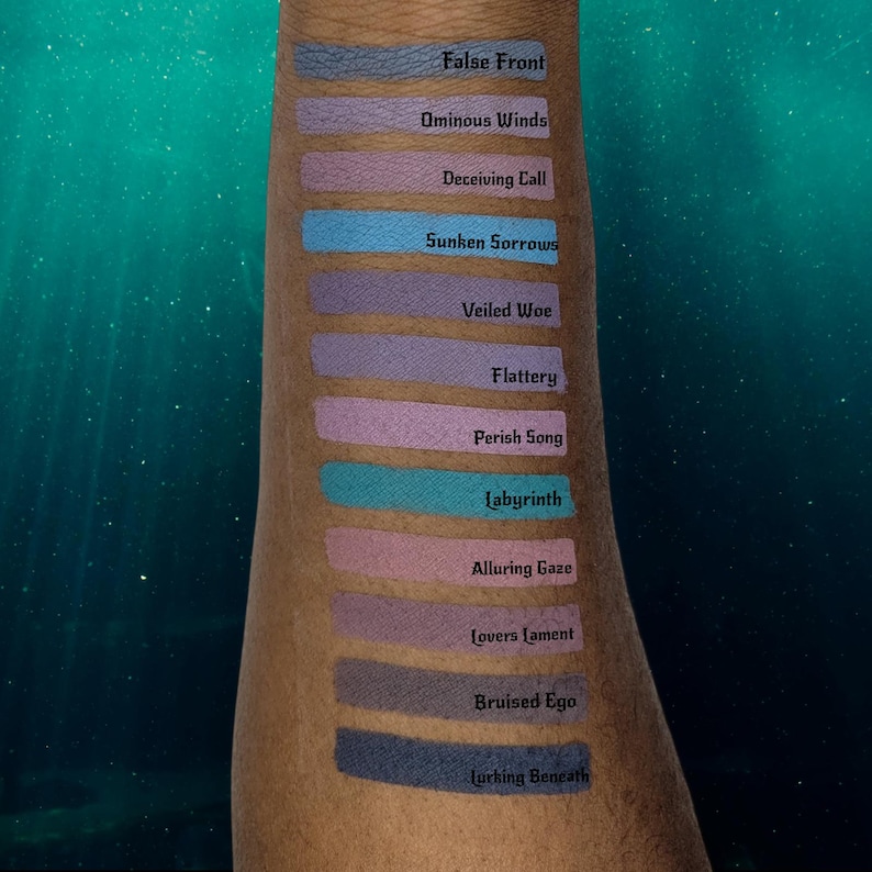 May include: A series of eyeshadow swatches in various shades of blue, purple, and pink are displayed on an arm. Each swatch has a name printed above it, including "False Front," "Ominous Winds," and "Sunken Sorrows." The background is a teal gradient.