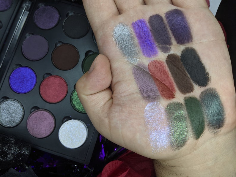 Op de afbeelding: Een zwart oogschaduwpalet met 15 verschillende tinten oogschaduw. De tinten vari&euml;ren van licht tot donker en omvatten glinsterende, matte en metallic afwerkingen. De oogschaduw is geswatcht op een hand om de kleur en textuur te laten zien.