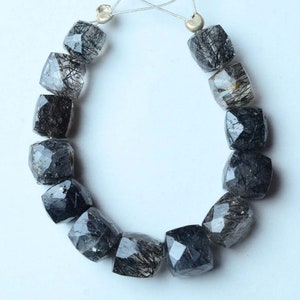 Könnte beinhalten: Eine Kette aus schwarzen Turmalin-Perlen, jede Perle ist facettiert und hat eine quadratische Form. Die Perlen sind auf einer durchsichtigen Schnur aufgefädelt und bereit für die Schmuckherstellung.