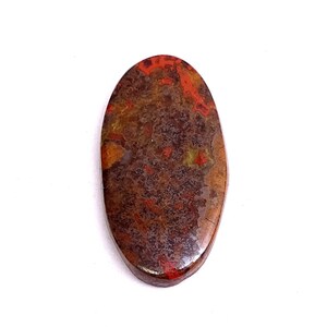 Puede incluir: Una gema de forma ovalada con una superficie pulida. La piedra presenta una mezcla de colores, incluyendo rojo, marrón y amarillo, creando un patrón moteado. La gema está sobre un fondo blanco.
