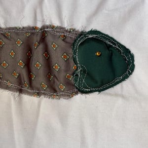 Op de afbeelding: Een wit T-shirt met een groene en bruine stoffen vis-applicatie. De vis heeft een gouden knoop-oog.