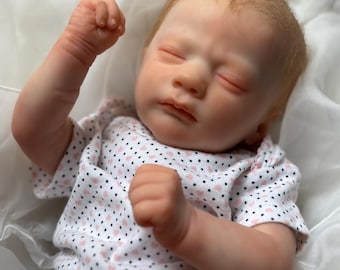 May include: A lifelike baby doll with closed eyes and a serene expression. The doll has fair skin, reddish-blonde hair, and is dressed in a white onesie patterned with small pink hearts. The doll's arms are raised, with fists clenched.