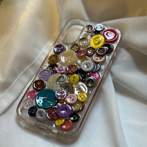 May include: Clear phone case decorated with an assortment of colorful buttons. The buttons vary in size, shape, and color, including red, yellow, pink, and blue. The case is set against a white fabric background.