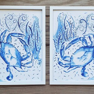 May include: Two framed watercolor prints featuring blue crabs in a stylized underwater scene. The artwork showcases the crabs amidst aquatic plants and swirling patterns, all rendered in shades of blue against a white background. The frames are white.