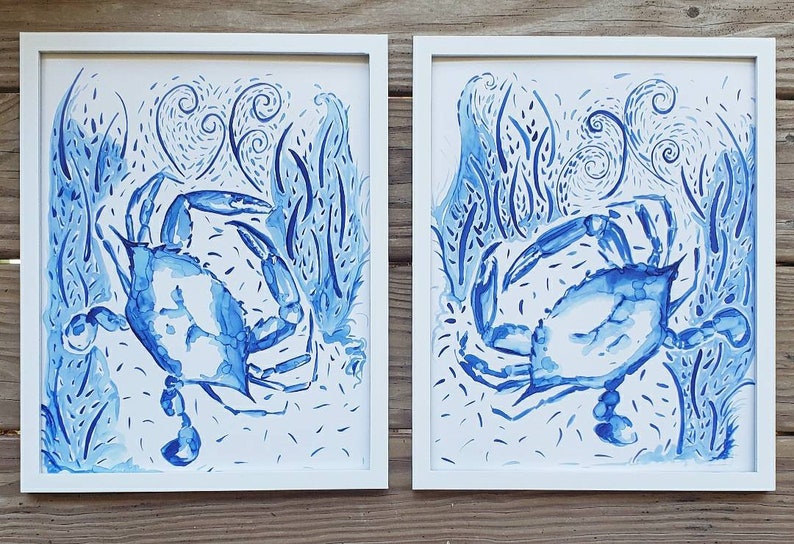 May include: Two framed watercolor prints featuring blue crabs in a stylized underwater scene. The artwork showcases the crabs amidst aquatic plants and swirling patterns, all rendered in shades of blue against a white background. The frames are white.