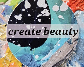 May include: A round sticker with the words "create beauty" in a gray band. The sticker has a colorful abstract design with blue, black, and white elements. The background features a palette of mixed paint colors.