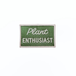 May include: A rectangular embroidered patch with a dark green background and a light-colored border. The word Plant is stitched in a white cursive script above the word ENTHUSIAST, which is stitched in white capital block letters.