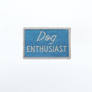 Peut inclure: Un écusson rectangulaire brodé avec un fond bleu et une bordure gris clair. Le mot "Dog" est brodé en lettres cursives blanches au-dessus du mot "ENTHUSIAST" qui est brodé en lettres capitales blanches en bloc.