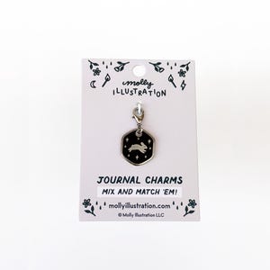 May include: A silver-toned hexagonal charm featuring a black enamel inlay with a white rabbit and star design. The charm is attached to a white display card printed with the text Molly Illustration, Journal Charms, Mix and Match 'Em!