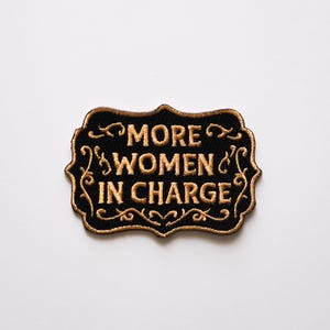 May include: Black and gold embroidered patch with the text "More Women in Charge" in a decorative font.
