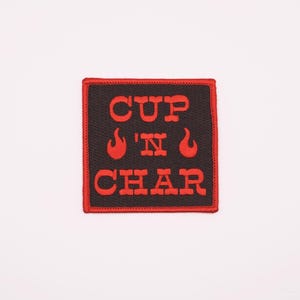 Puede incluir: Un parche bordado negro y rojo con el texto "CUP 'N CHAR" y dos diseños de llamas.