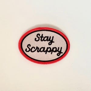 May include: Oval-shaped embroidered patch with the words "Stay Scrappy" in black script. The patch has a white background with a red border.