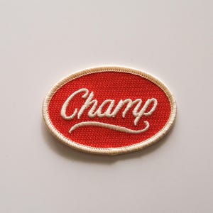Puede incluir: Parche ovalado rojo con la palabra "Champ" bordada en escritura blanca. El parche tiene un borde color crema y una superficie texturizada. Adecuado para chaquetas, bolsos u otros artículos de tela. Mide unos 7 cm de largo.