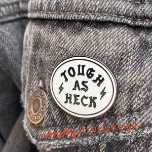 May include: White enamel pin with a black outline and the text "Tough as Heck" with lightning bolts on either side.