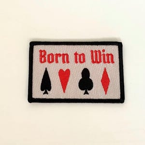 Puede incluir: Parche bordado con el texto "Born to Win" en rojo, con los palos de la baraja: picas, corazones, tréboles y diamantes. El parche tiene un fondo gris claro y un borde negro.