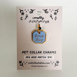 May include: A light pink pet collar charm tag with a gold border and a light blue centre. The tag reads "Love Bug" in white text. The tag is attached to a light pink card with the text "PET COLLAR CHARMS" and "MIX AND MATCH 'EM!"