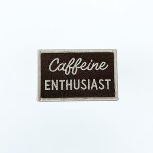 May include: A rectangular embroidered patch with a dark brown background and a light beige border. The text "Caffeine" is stitched in a white cursive script font above the word "ENTHUSIAST" which is stitched in a white block font.