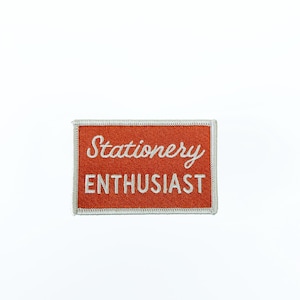 May include: A rectangular embroidered patch with a burnt orange background and a white border. The word Stationery is written in a white cursive script above the word ENTHUSIAST, which is written in white capital block letters.