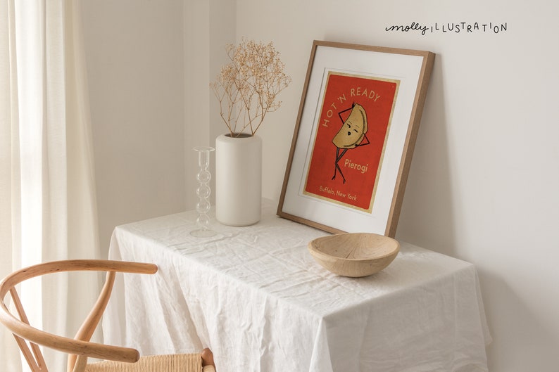 May include: A framed illustration of a pierogi with a face and arms, with the text "I'm ready, Hot'n Pierogi, Buffalo, New York". The illustration is on a red background and the frame is light wood.