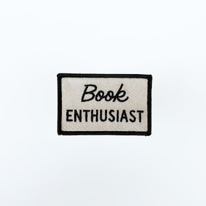 May include: A rectangular embroidered patch with a light beige background and a thick black border. The word Book is stitched in a black cursive script font above the word ENTHUSIAST, which is stitched in a bold black sans-serif font.