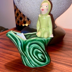 May include: A vintage ceramic planter with a green tree trunk design. A small figurine of a seated figure in a green outfit with a hood is perched on the planter's edge. The figurine has a painted face with red lips and green eyes.