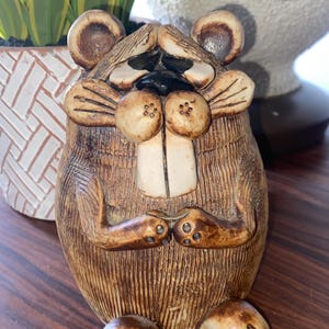 Puede incluir: Una figurita de cerámica de un animal marrón y blanco con orejas grandes, una boca ancha y una superficie texturizada. El animal está sentado con las patas cruzadas.