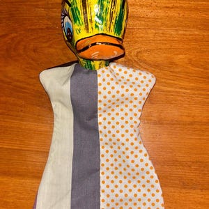 Op de afbeelding: Een handpop met een eendenkop met een geel, groen en bruin geschilderd ontwerp. De pop heeft een stoffen lijfje met verticale strepen van lichtgroen, grijs en een witte stof met oranje stippen.