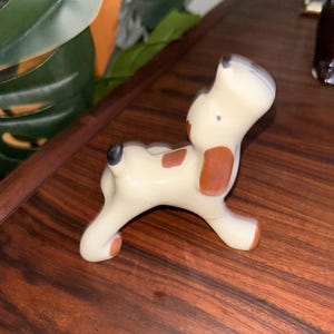 Puede incluir: Figura de perro de cerámica color crema en pose juguetona, con manchas y detalles marrones. El perro tiene una nariz negra y una mancha gris en la cabeza. Colocada sobre una superficie de madera oscura.