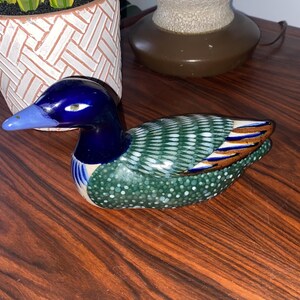 May include: A ceramic duck figurine with blue, green, white, and brown paint. The duck has a blue head and neck, green and white wings, and a brown tail.