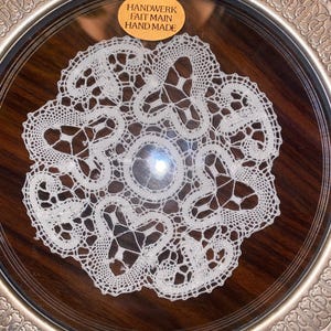 May include: A white crocheted doily with a delicate floral design, displayed on a silver tray with a wooden base. The doily is labeled "Handwerk Fait Main Hand Made".