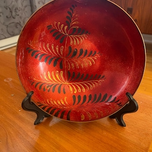 Peut inclure: Un bol décoratif rouge avec un bord doré et un support noir. Le bol présente un motif de feuilles dorées et noires. Le bol est exposé sur une surface en bois.