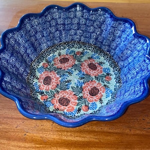 Puede incluir: Un cuenco de cerámica azul con borde festoneado. El cuenco presenta un diseño floral en el centro con flores rojas y rosas sobre un fondo azul claro. El borde exterior tiene un patrón azul detallado.