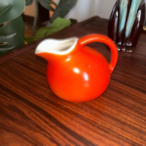 May include: A small, round, orange ceramic creamer with a handle and a white interior. The creamer is on a dark wood surface. A decorative vase is in the background.