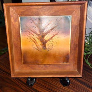 May include: A framed artwork featuring a tree silhouette against a sunset backdrop. The artwork is in a square format, set within a wooden frame. The color palette includes warm tones of orange, brown, and yellow, with a touch of blue.