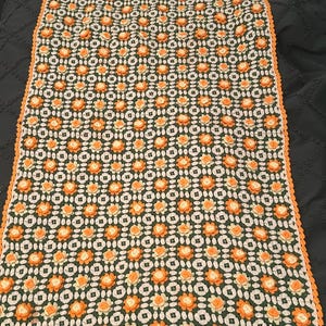 May include: A rectangular, handmade crochet blanket featuring a repeating floral pattern in orange and white against a dark green background. The blanket has an orange border and is likely used as a decorative throw.