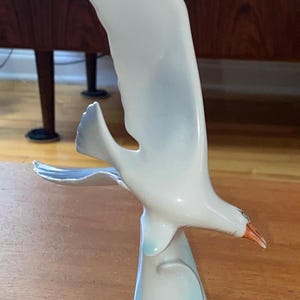 Könnte beinhalten: Eine Keramikskulptur einer Möwe im Flug. Der Vogel ist überwiegend weiß mit dezenten blauen und grauen Akzenten auf den Flügeln und am Sockel. Der Schnabel ist hellorange. Die Skulptur steht auf einer Holzoberfläche.