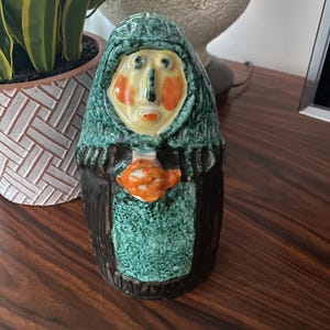 May include: A ceramic figurine of a person wearing a green hooded robe and holding a small orange object. The figurine has a stylized face with orange cheeks and a yellow nose.