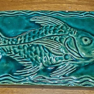 May include: A rectangular, teal-colored ceramic tile featuring a detailed relief of a fish. The fish is centered, with fins and scales clearly defined. Wavy lines border the tile, adding a decorative element. The tile is likely for home decor.