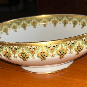 Puede incluir: Cuenco de cerámica blanca de forma ovalada con borde y base dorados. El exterior del cuenco presenta un patrón decorativo de diseños florales verdes, dorados y rojos. El cuenco está sobre una superficie de madera.