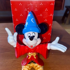 K&ouml;nnte beinhalten: Eine Keramikfigur von Mickey Mouse als Zauberer, mit einem blauen Hut mit Sternen und einem Mond, und einem roten Umhang. Die Figur hat wei&szlig;e Handschuhe und gelbe Schuhe. Die rote Schachtel dahinter tr&auml;gt das Wort "Schmid".
