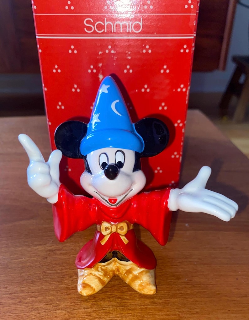 K&ouml;nnte beinhalten: Eine Keramikfigur von Mickey Mouse als Zauberer, mit einem blauen Hut mit Sternen und einem Mond, und einem roten Umhang. Die Figur hat wei&szlig;e Handschuhe und gelbe Schuhe. Die rote Schachtel dahinter tr&auml;gt das Wort "Schmid".