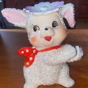 Puede incluir: Una figurita de cerámica blanca de un cordero con un sombrero de flor morado y una pajarita roja con lunares blancos. El cordero está cubierto de purpurina blanca.