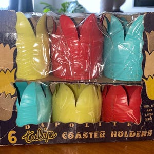 May include: A vintage box containing six tulip-shaped coaster holders in yellow, red, and teal. The box is labelled "Dapolite" and "6 tulip coaster holders." The box is brown with tulip illustrations and the text "Will fit any large glass."