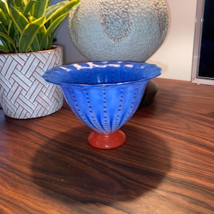 May include: A blue glass bowl with a red base. The bowl has a flared rim and is decorated with small, evenly spaced red dots. The bowl is sitting on a dark wood surface.