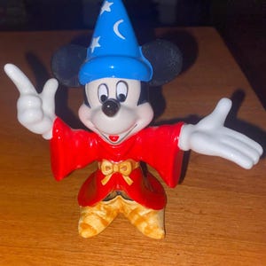 K&ouml;nnte beinhalten: Eine Mickey Mouse-Figur als Zauberer. Mickey tr&auml;gt einen blauen Hut mit wei&szlig;en Sternen und einem Mond, einen roten Umhang und eine goldene Hose. Seine wei&szlig; behandschuhten H&auml;nde sind ausgestreckt.