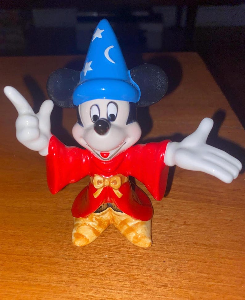 K&ouml;nnte beinhalten: Eine Mickey Mouse-Figur als Zauberer. Mickey tr&auml;gt einen blauen Hut mit wei&szlig;en Sternen und einem Mond, einen roten Umhang und eine goldene Hose. Seine wei&szlig; behandschuhten H&auml;nde sind ausgestreckt.